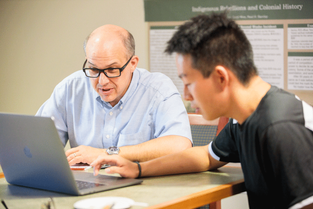 Professor Paul Barclay sits with a student at a laptop.