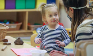 A young child looks at the childcare worker helping her