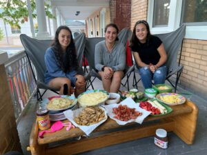 Rachel Hurley’23, Anna Radcliff’23 and Kellie Kottman’23 sit on a couch behind a table filled with food at the Global Foods House in the Monroe Neighbhorhood.