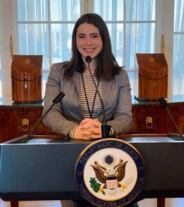 Kaitlin Ahern sits at a desk with the United States Department State seal in front