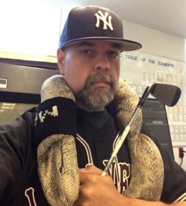 Chip Nataro wears a Yankees baseball cap and Lafayette baseball jersey while carrying a golf club and having what appears to be faux snakeskin draped over his shoulders