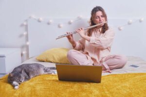 A woman plays the flute on her bed facing her laptop with her cat sitting nearby