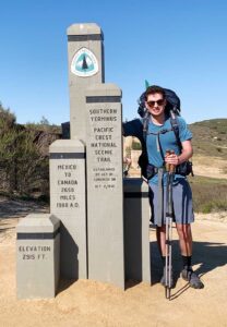 Ben Smaruk stands at Pacific Coast Trail trail sign in Mexico wearing hiking gear