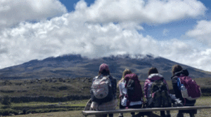 Four students sit on a bench and look at a mountain as clouds are above.