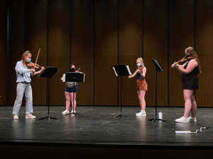 Four violinists rehearse on stage while wearing masks.