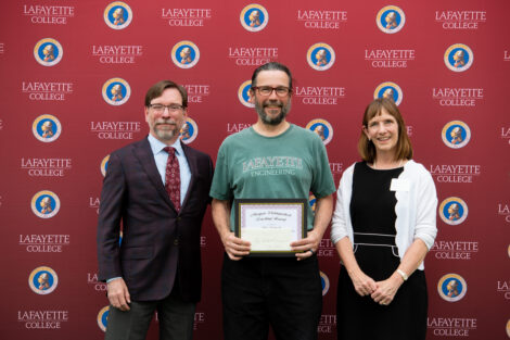 An employee holding his award poses in front of the Lafayette-branded backdrop at the conclusion of the faculty-staff awards event at Fisher Stadium