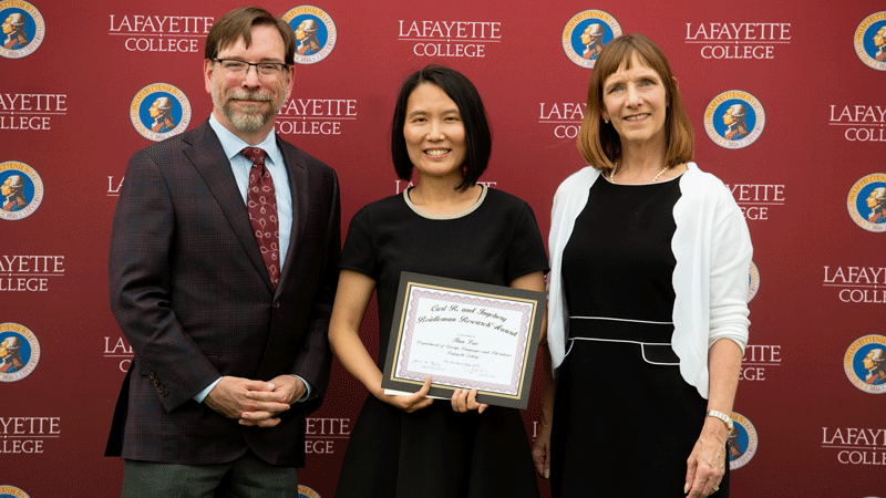 Han Luo, assistant professor of foreign languages and literatures, holds an award while flanked by Provost John Meier and President Alison Byerly in front of a Lafayette-branded backdrop at Fisher Stadium.