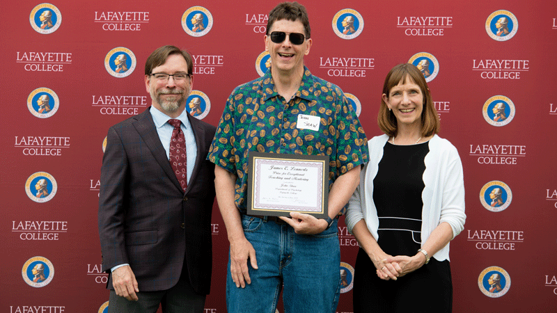 John Shaw holds his award while flanked by Provost John Meier and President Alison Byerly in front of a Lafayette-branded backdrop at Fisher Stadium.