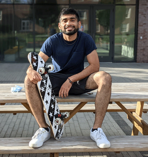 Ali Sulta Sikander sits on a wooden bench while holding a black and white skateboard.