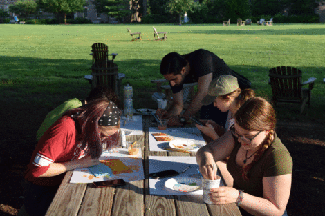 Four students paint cacti while seated at a picnic table near the Quad.