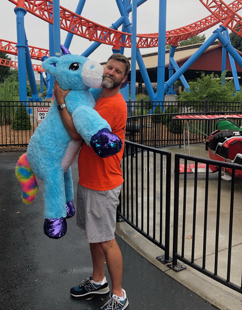 Professor Trent Gaugler holds a blue unicorn stuffed animal as if it's kissing him on the cheek.