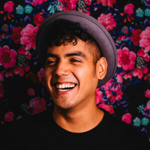 Saul Flores smiles while wearing a hat, with a backdrop of painted flowers