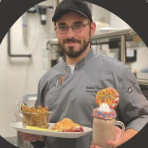 Chef Andrew Hershbine carries a tray with two food items in one hand and a milkshake with a cookie on top in the other hand.