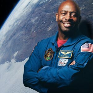 Leland Melvin wears a blue NASA jacket in front of an image of the earth taken from outer space