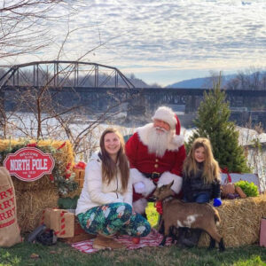 A man in a Santa Claus costume sits with two girls and a young goat for a photo