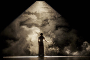A darkened stage shows a woman silhouetted with clouds in the background.