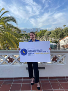 Shiloh Harrill stands with a poster reading 21st Undergraduate Research Conference on the European Union in California, after presenting her research paper.
