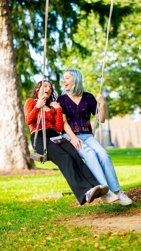 Two students laughing while sitting on a swing together on a sunny day.
