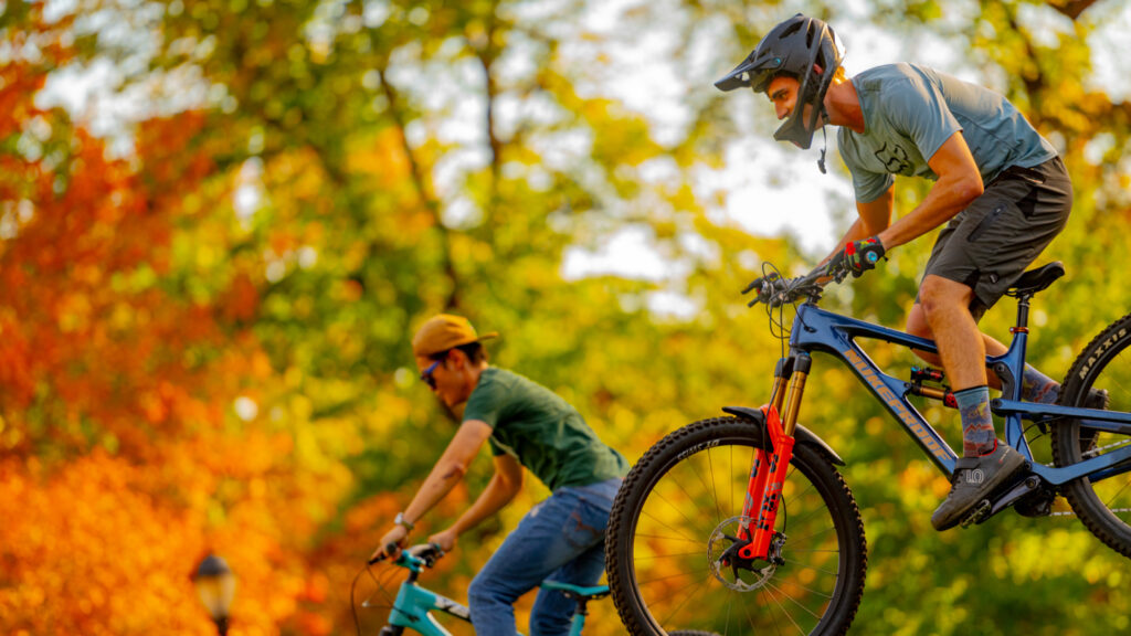 Action shot from the side of two students riding bikes with fall foliage around them.