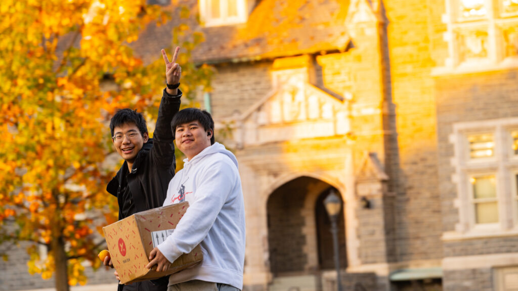 Two students smiling in front of Hogg Hall on a sunny day. The person on the left is holding up a peace sign while the person on the right carries a cardboard box.