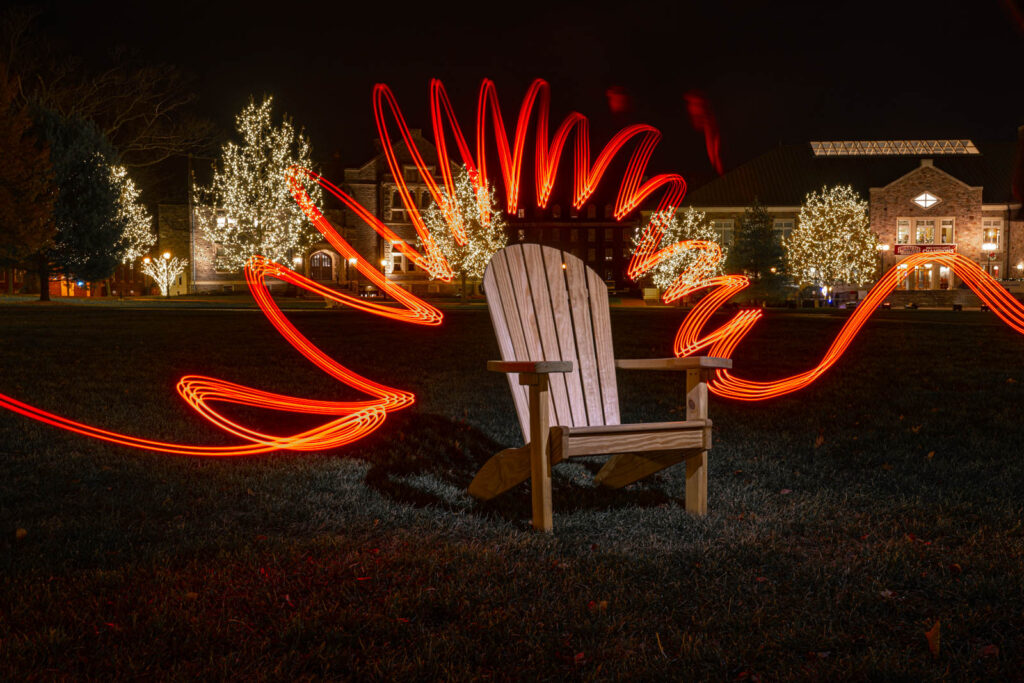A night scene of an adirondack chair. The chair has been accentuated with light-painting, creating the effect of a red starburst around the chair. In the background twinkle lights are visible. 
