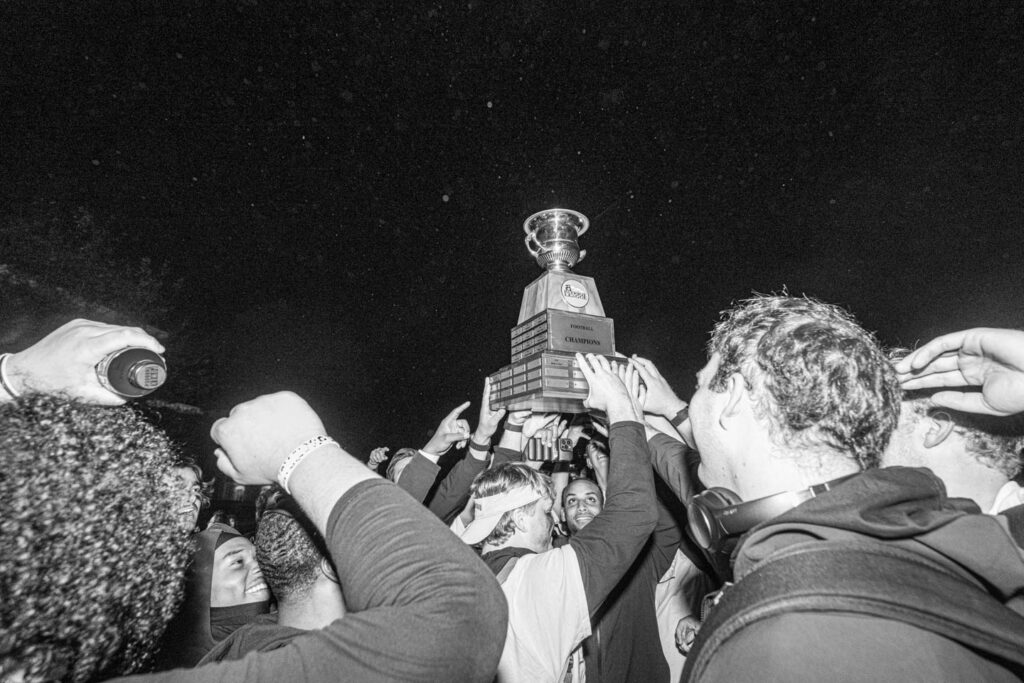 A black and white photo of a group of male football players hoisting the patriot league championship trophy abover their heads. 