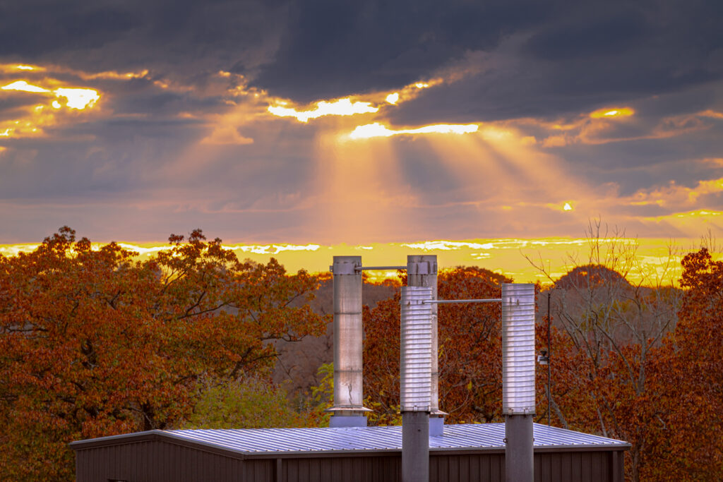 Three metal chimneys visibly emit heat. Sun breaks through the clouds in the background. 