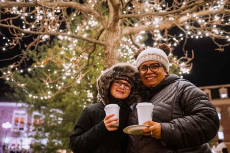 A staff member and student hold cocoa on the Quad with a tree lit up in the background with twinkle lights.