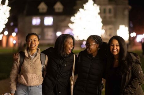 A group of four students laugh as the trees are lit up in the background on the quad.