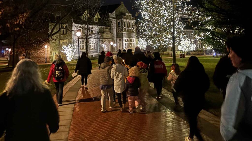 During Las Posadas a group of carolers walk through the Quad as the trees are lit with lights in the background.