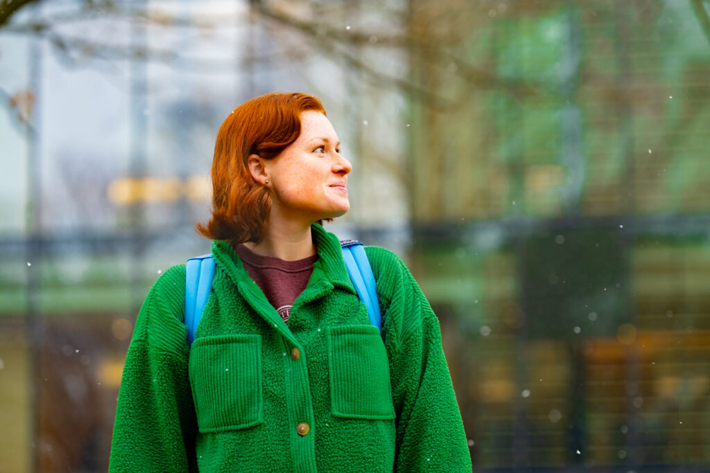 A student in a bright green jacket facing the camera looks to her left while snow falls. 