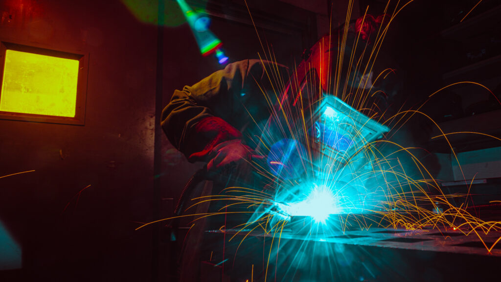 A welder uses their tools on a steel beam to create sparks of light.