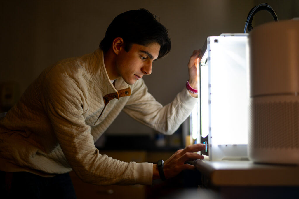 A student, in a dark room, leans in to calibrate a 3d printer. The printer is square shaped and emits a bright light. 
