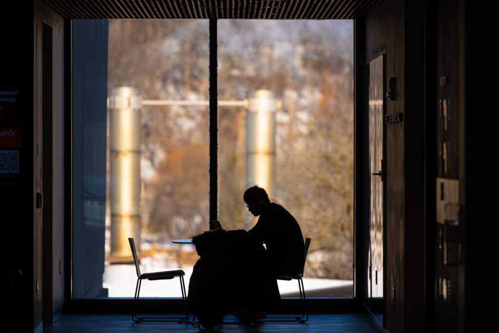 A silhouette of a student sitting at a table studying next to a large window.