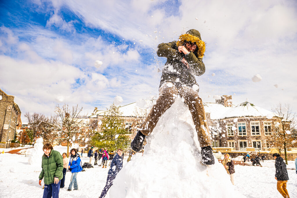A student in a parka sits on a mound of snow getting hit with snowballs.
