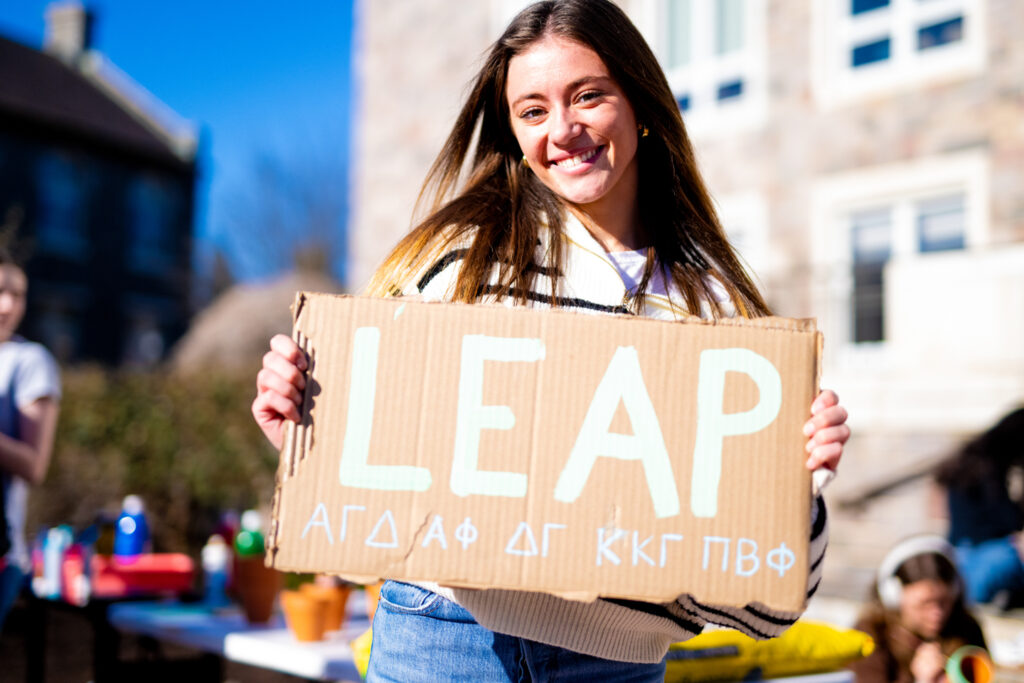 A student holds a cardboard sign that says LEAP and indicates the involvement of several Greek Life organizations. 