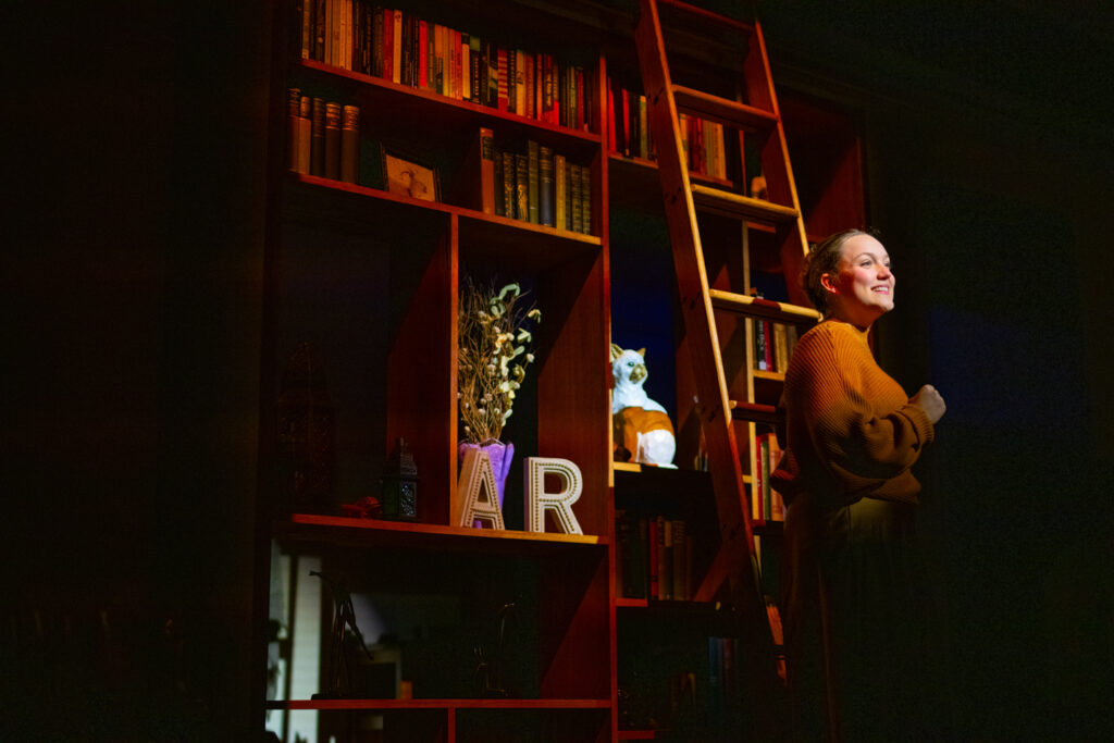 A person poses smiling in a spotlight on stage with bookshelves behind them.