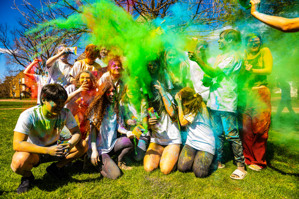 A group of people smile at the camera as green abd blue powder is thrown all over them.