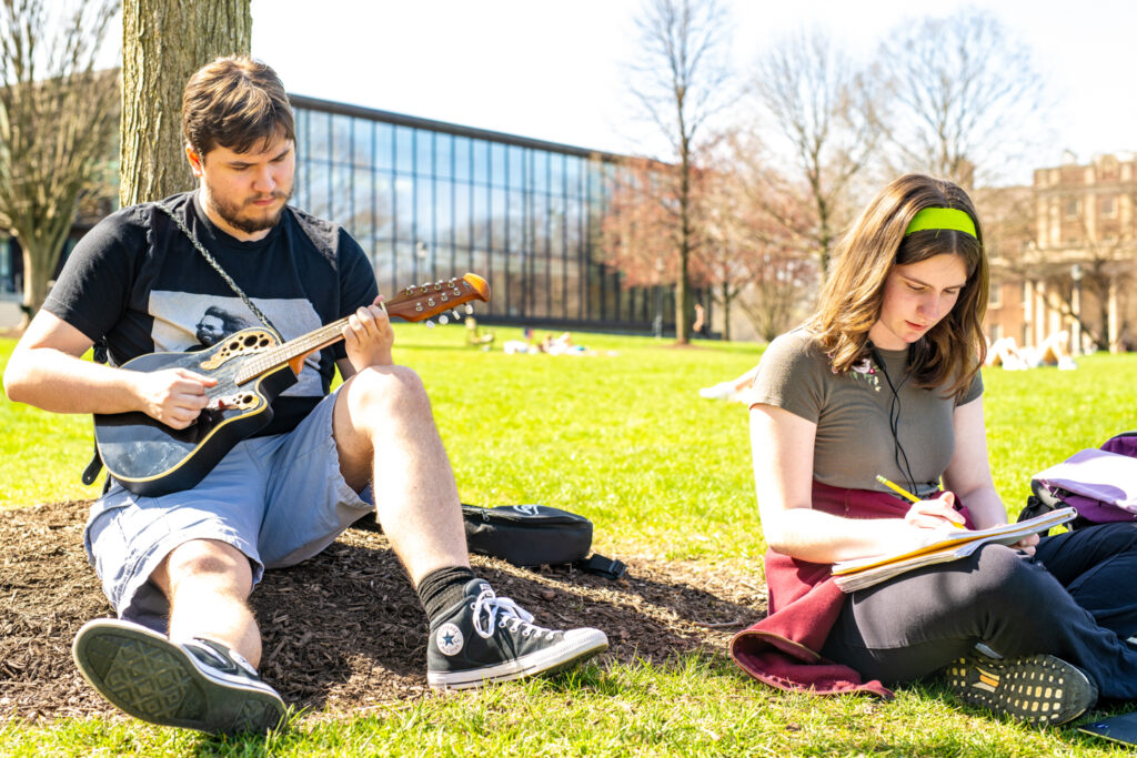 One person leans against a tree playing guitar while another person sits next to them writing in a journal.