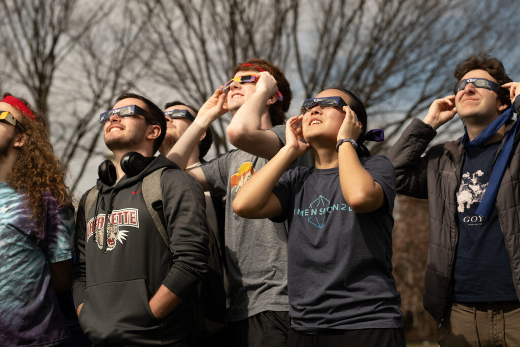 A group of people wearing Eclipse safety glasses look up towards the sky.