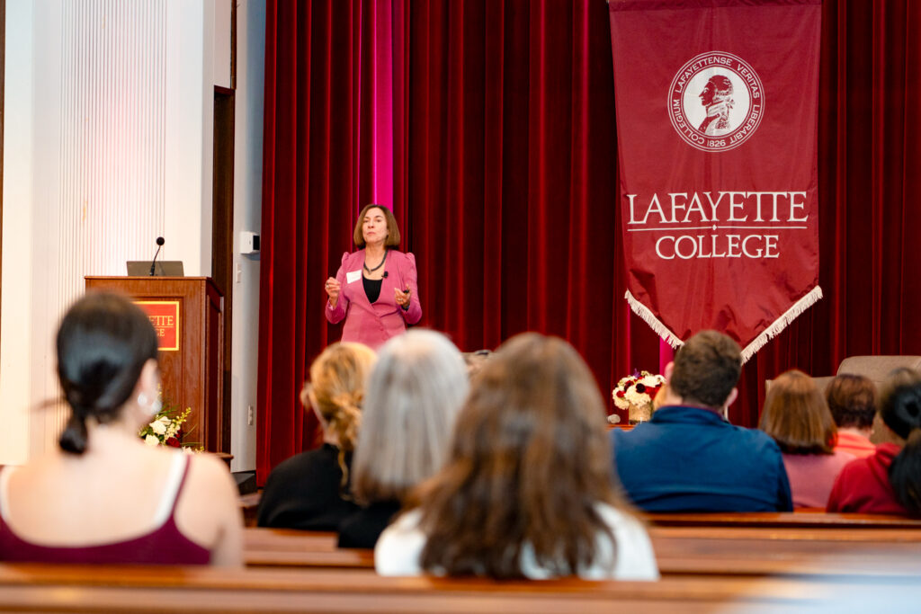 A woman stands on a stage, in front of an audience, with a red curtain. A flag hangs from the curtain. The flag reads, "Lafayette College."