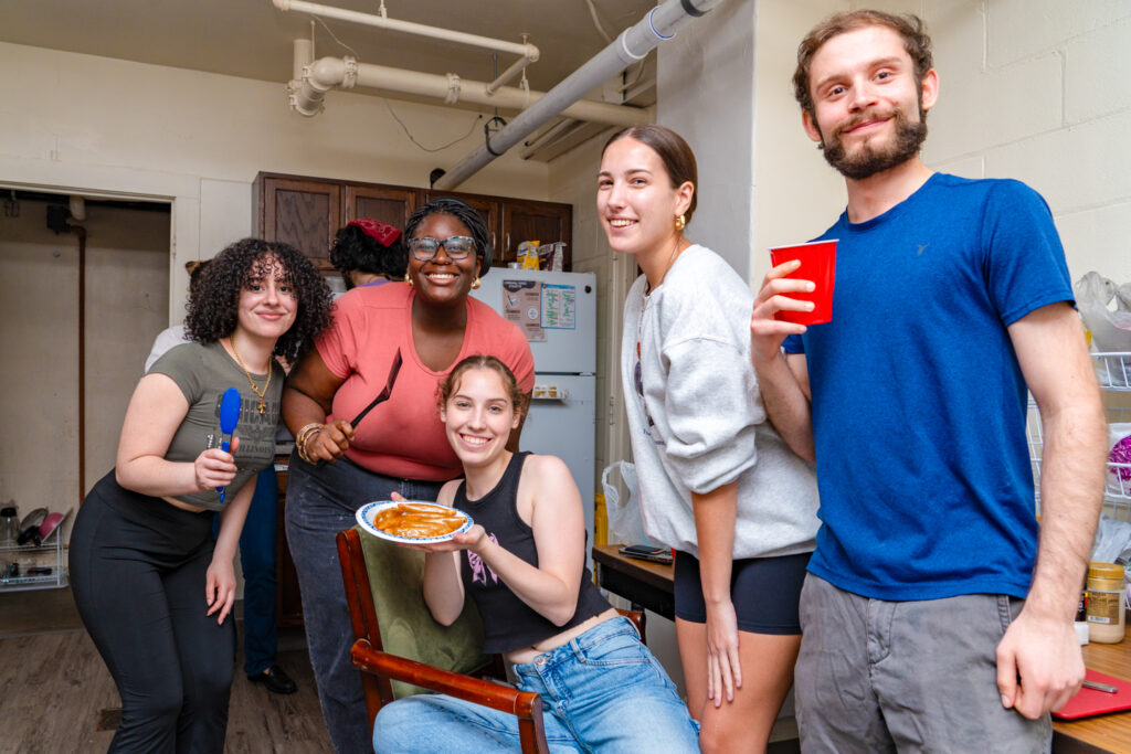 five students stand in a basement kitchen with white walls, and pipes in the ceiling. A girl in the middle of the group is holding a plate of food. 