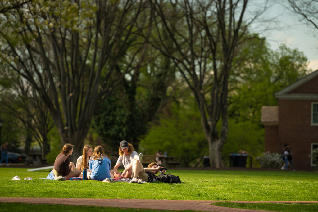 A group of students sit on the green grass of Lafayette College's Quad. They are all looking at the camera. 