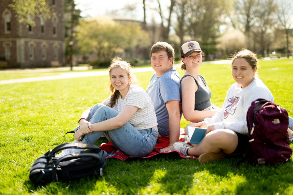 Four students sit on the green grass of Lafayette College's Quad. They are all looking at the camera. 