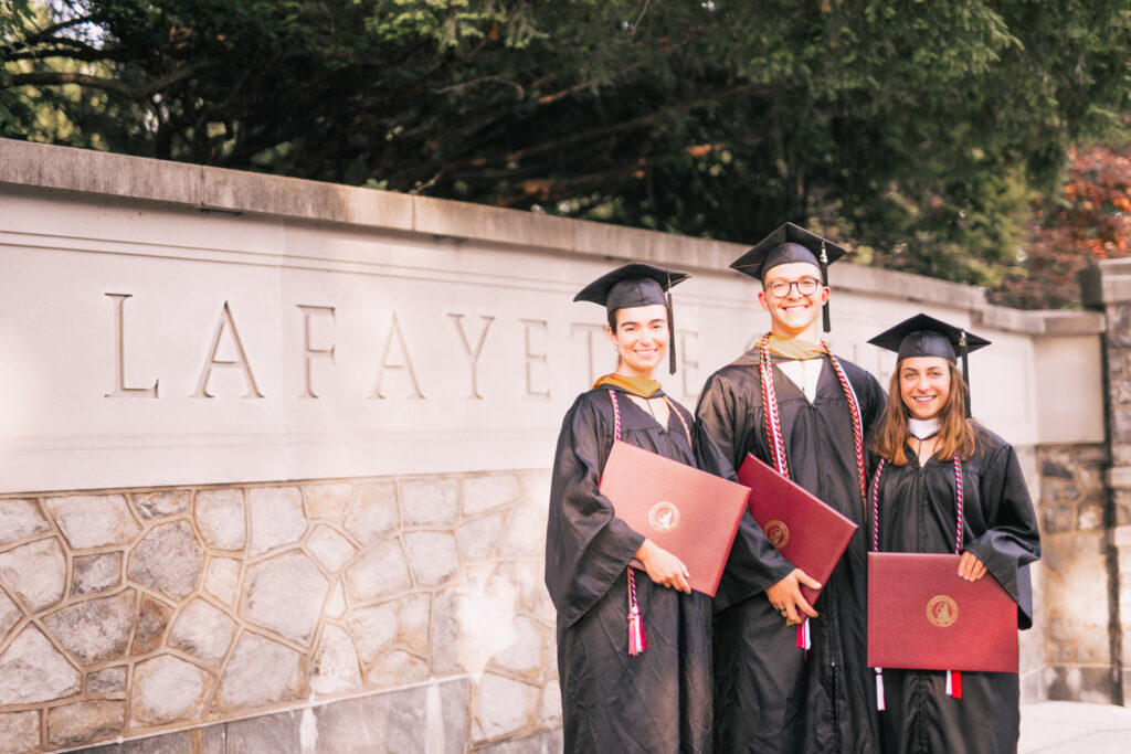 3 People in graduation clothing stand in front of the "Lafayette College" sign holding their degrees.