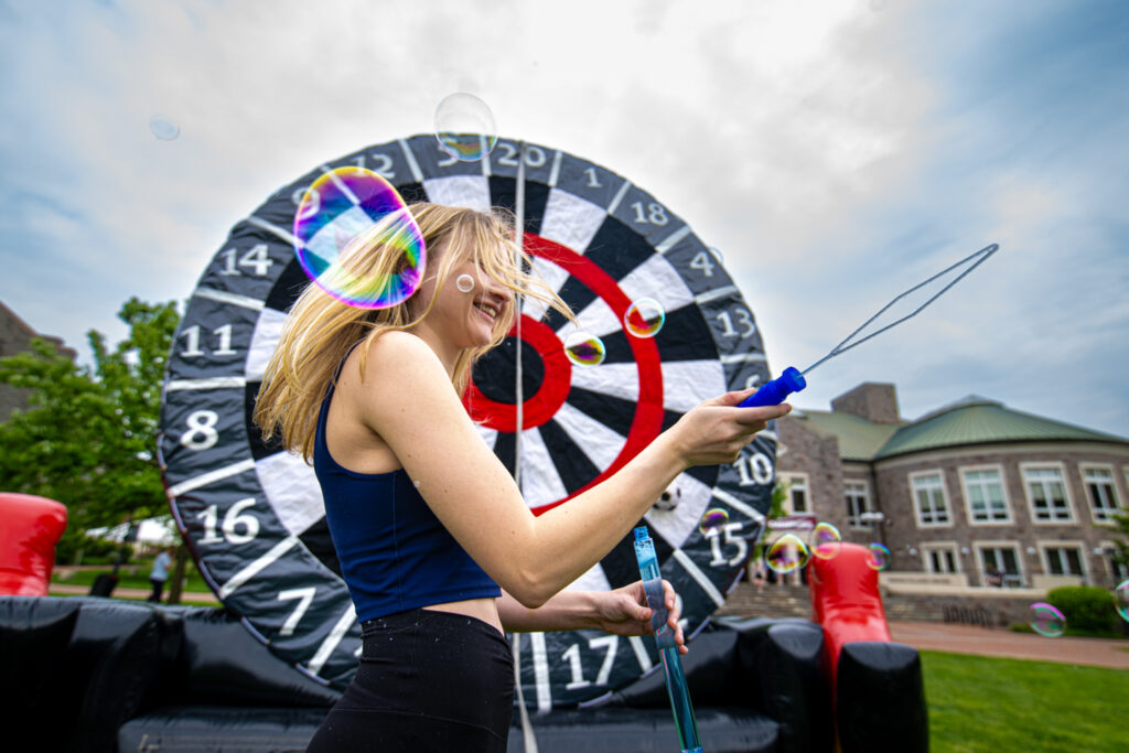 A young woman, in front of a large target, spins in circles producing large bubbles from a bubble wand. 