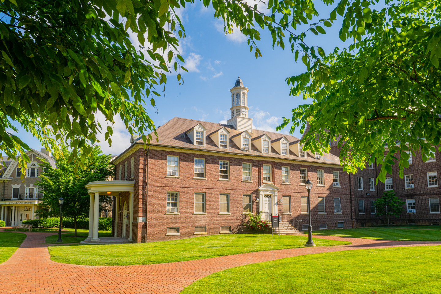 A brick building with a cupola is framed by green leaves and set against a blue sky. In front of the building is a brick path and grassy lawn. 