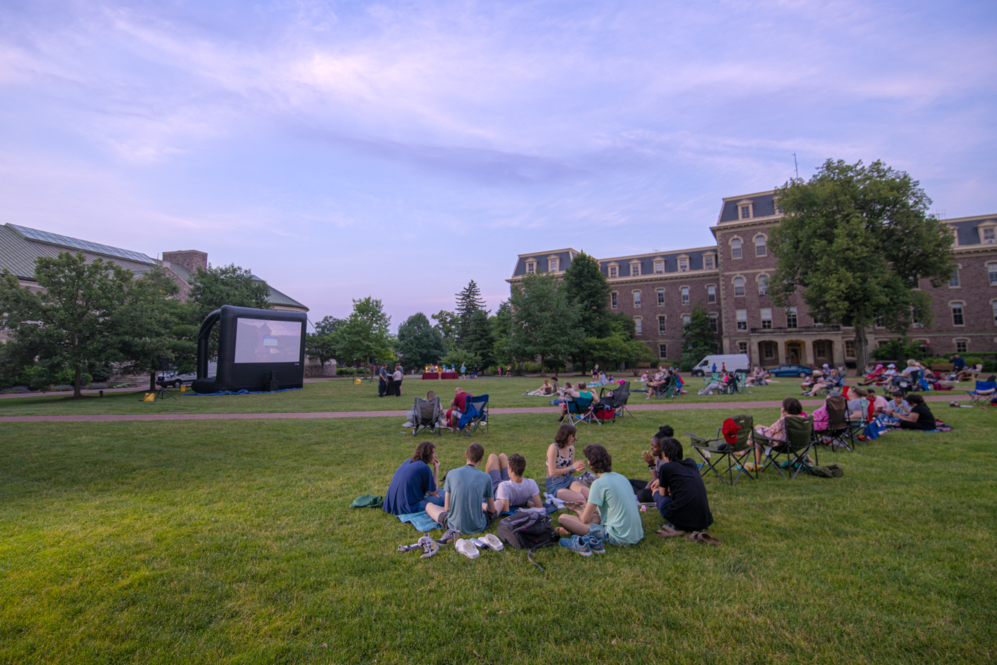 A group of people are aranged in a circle on the green grass of Lafayette College's Quad. Behind them are other members of the Easton Pa community. There are all waiting for a movie to be played on a screen, visible in the background. Pardee Hall can be seen in the background also. 