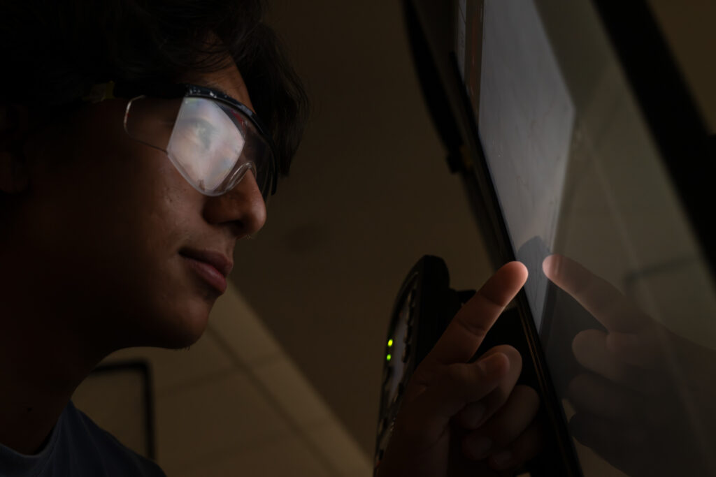 Close up photo of a person in safety glasses looking at a computer with the screen's reflection in their glasses.
