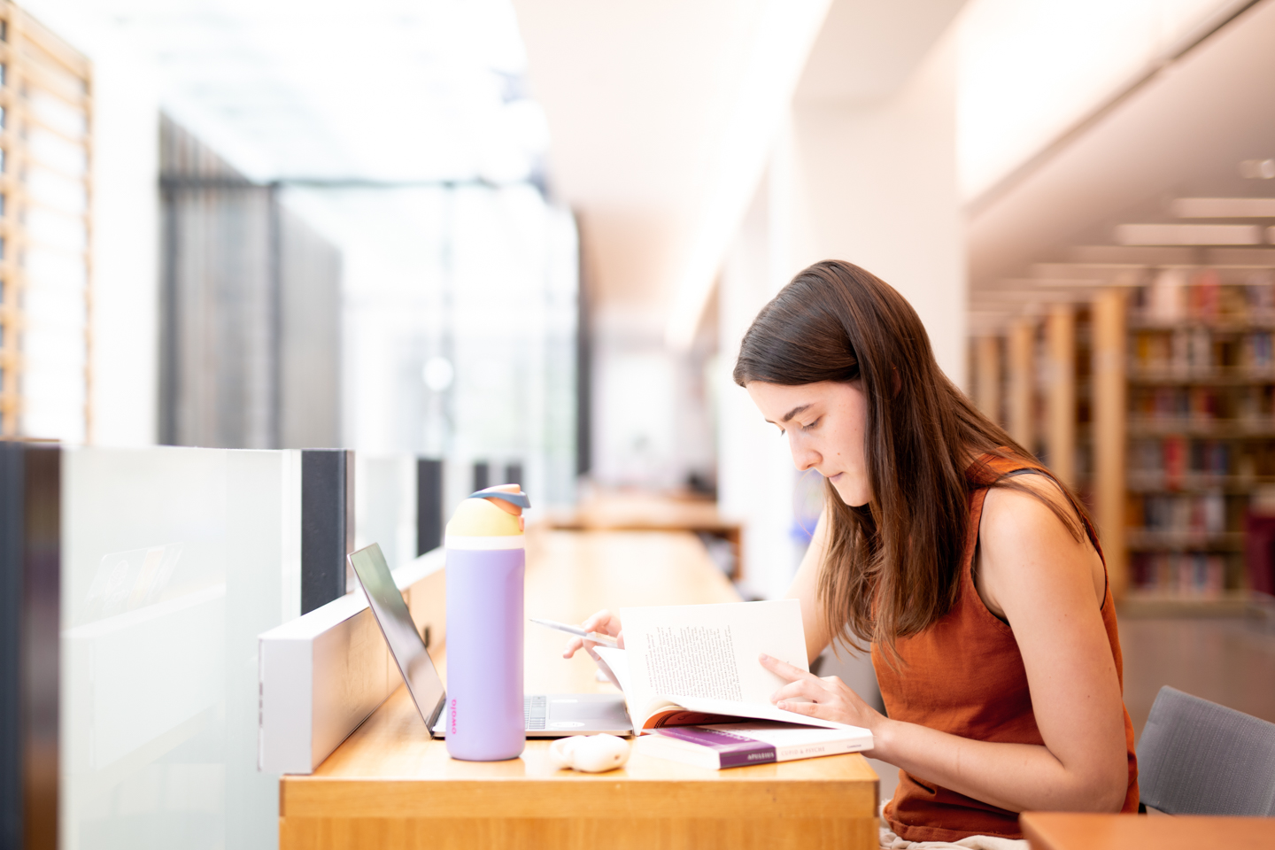 A female student wearing an red-orange sleeveless shirt is seated on the right side of the frame reading a book inside of a library. The student's laptop is open in front her and she has a purple thermal mug. 